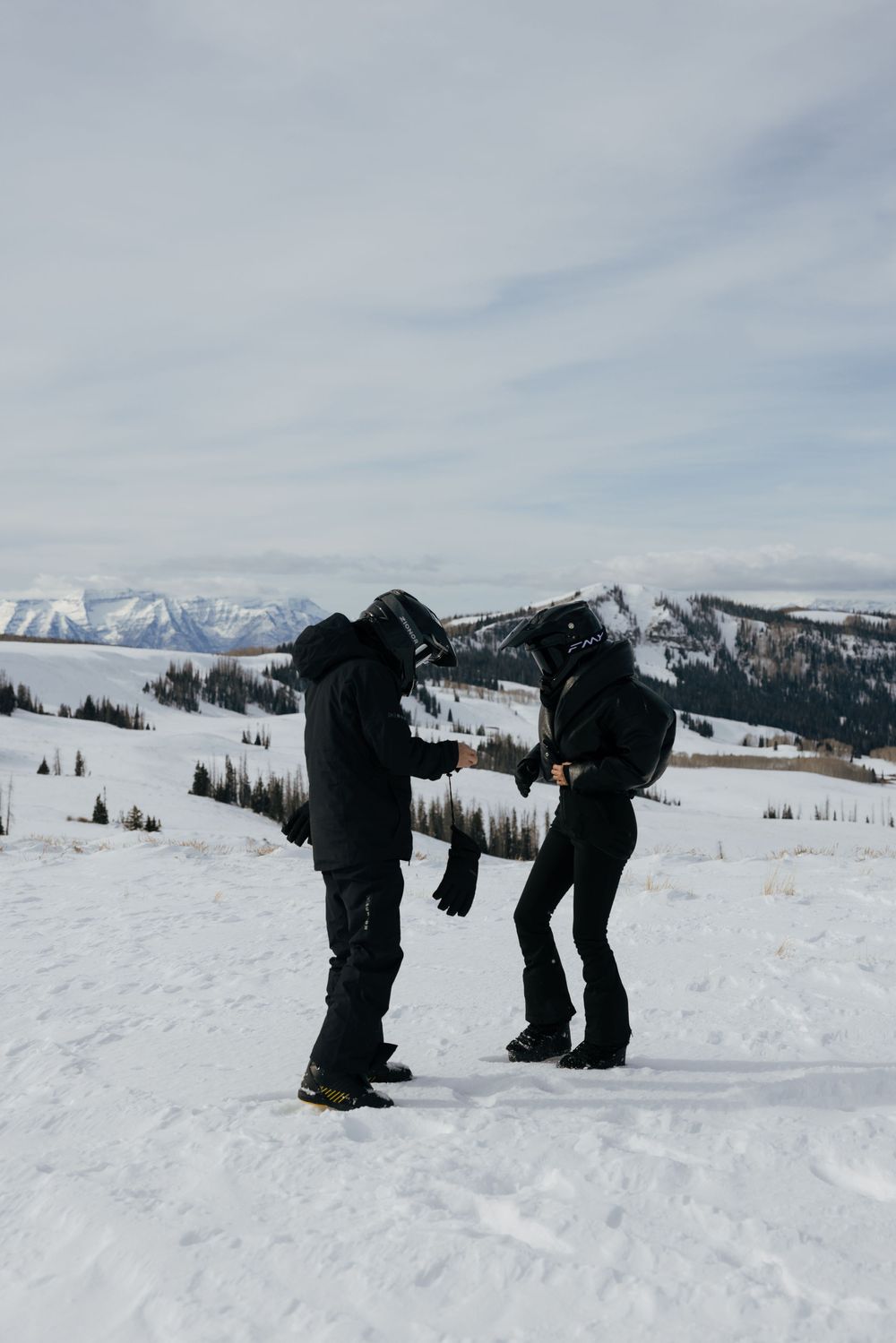 Two snowboarders playfully bow to each other on a snowy mountain slope with snow-capped peaks in the background.