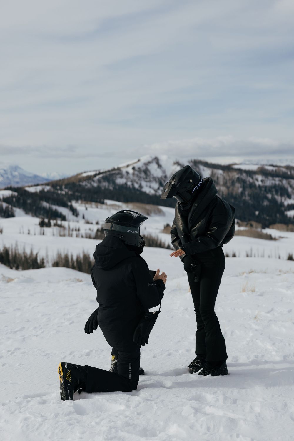 Two figures kneel in the snow against a backdrop of snow-covered mountains during what appears to be a proposal moment.