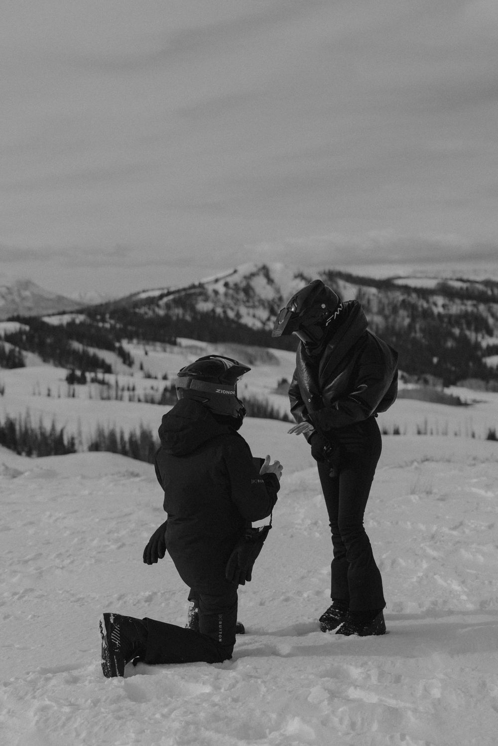 Two people kneel in the snow while skiing in a mountainous winter landscape with snow-covered peaks in the distance.