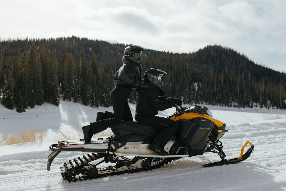 Snowmobiles racing across a snowy winter landscape with mountains and forest in the background.