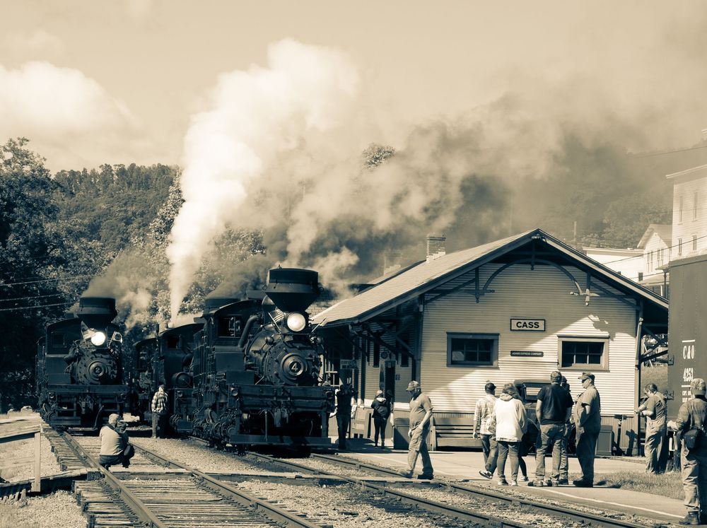 A vintage panoramic black and white photograph shows a steam locomotive and train station platform with passengers.