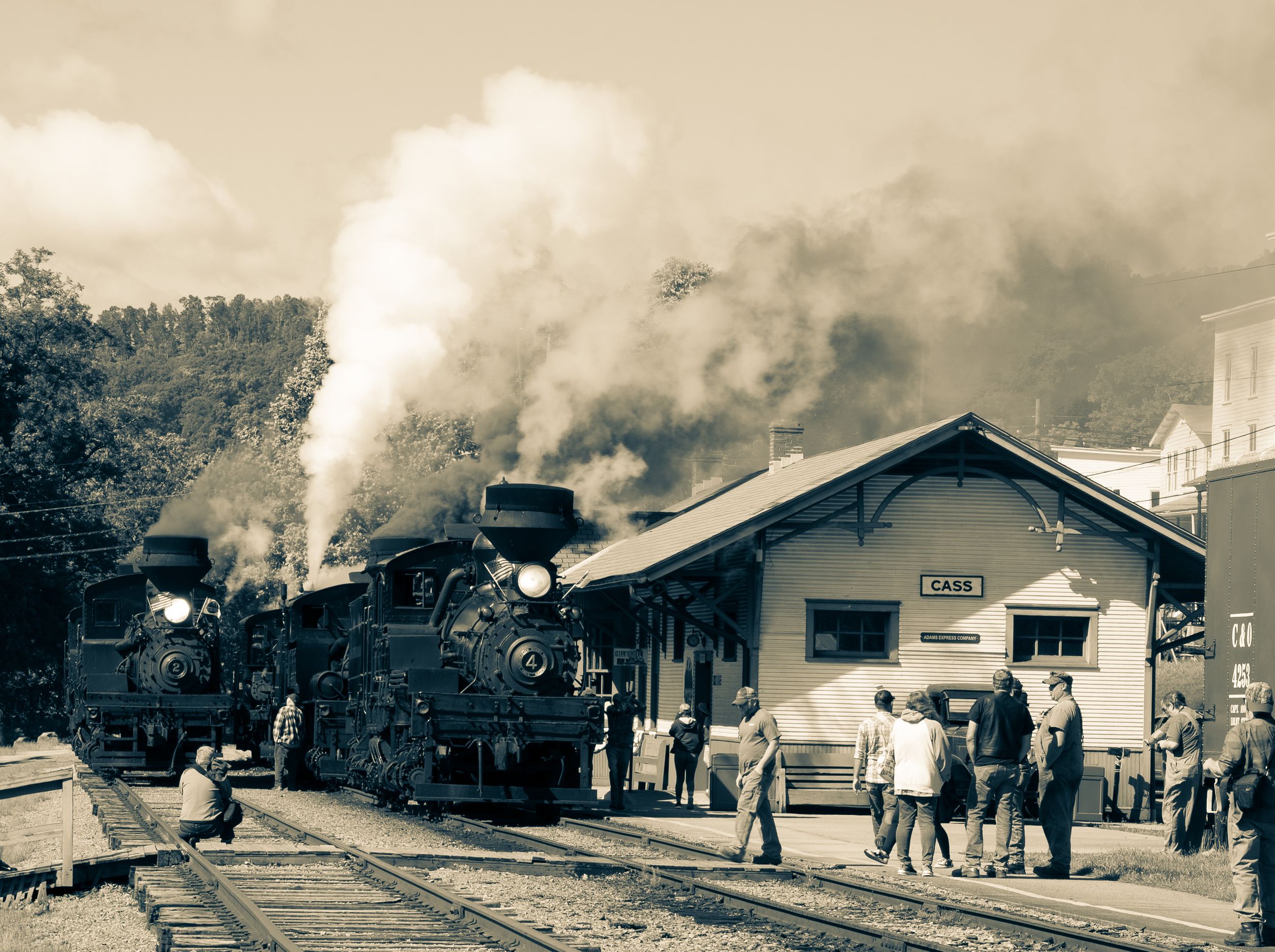 A vintage panoramic black and white photograph shows a steam locomotive and train station platform with passengers.