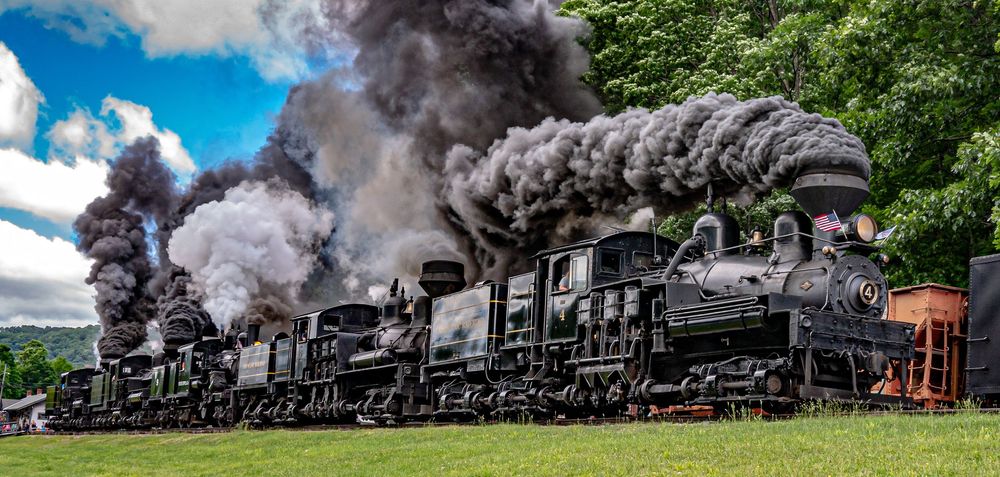 Multiple vintage steam locomotives emit dark smoke while operating on railroad tracks through green countryside.