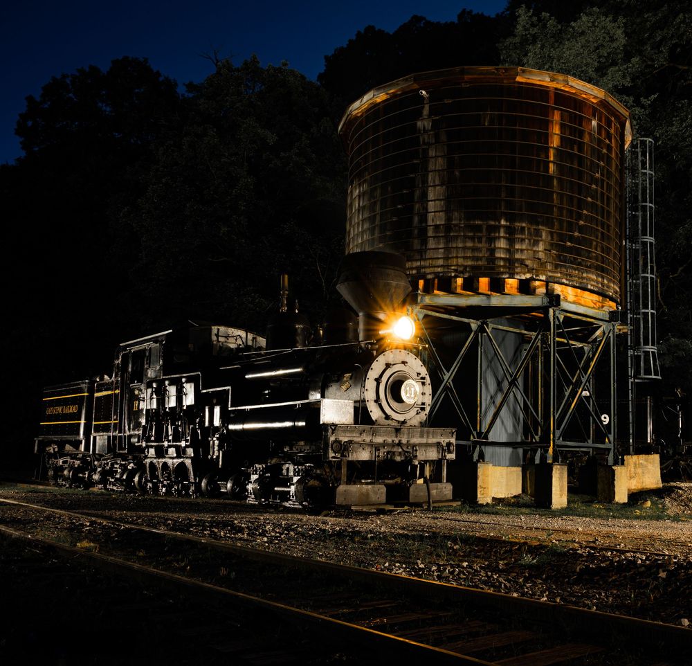 Nighttime industrial scene with illuminated train cars and storage silos casting dramatic shadows against dark surroundings.