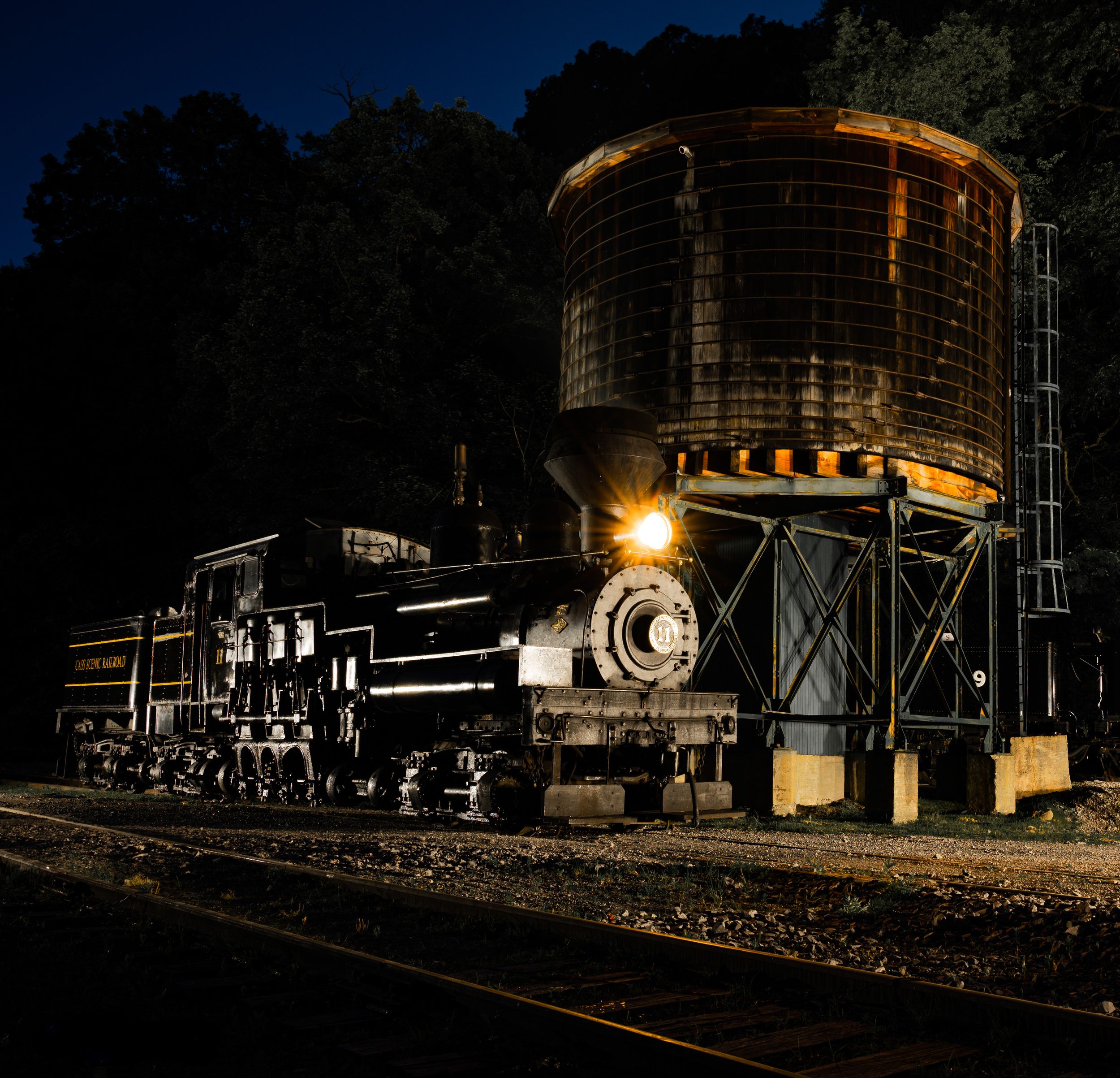 Nighttime industrial scene with illuminated train cars and storage silos casting dramatic shadows against dark surroundings.