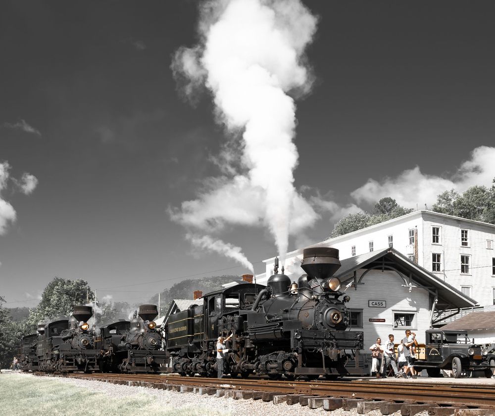 A black and white panoramic view of vintage steam locomotives lined up at a train station with billowing smoke.