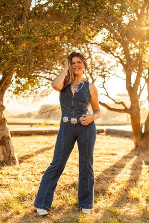 Person wearing denim jumpsuit poses in golden sunlit field with trees.