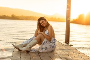 Person in floral dress relaxing on wooden dock during golden sunset over water.