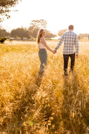 Couple walking hand in hand through a sun-lit meadow with golden grass.