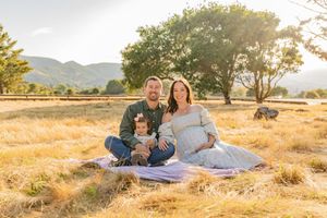 Family sitting on blanket in golden field during sunset with mountains and trees in background.