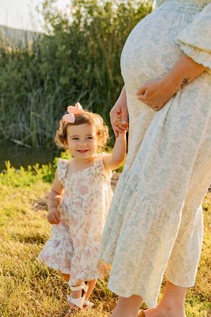Little girl in a floral dress plays in a sunlit meadow during golden hour as someone stands nearby watching.