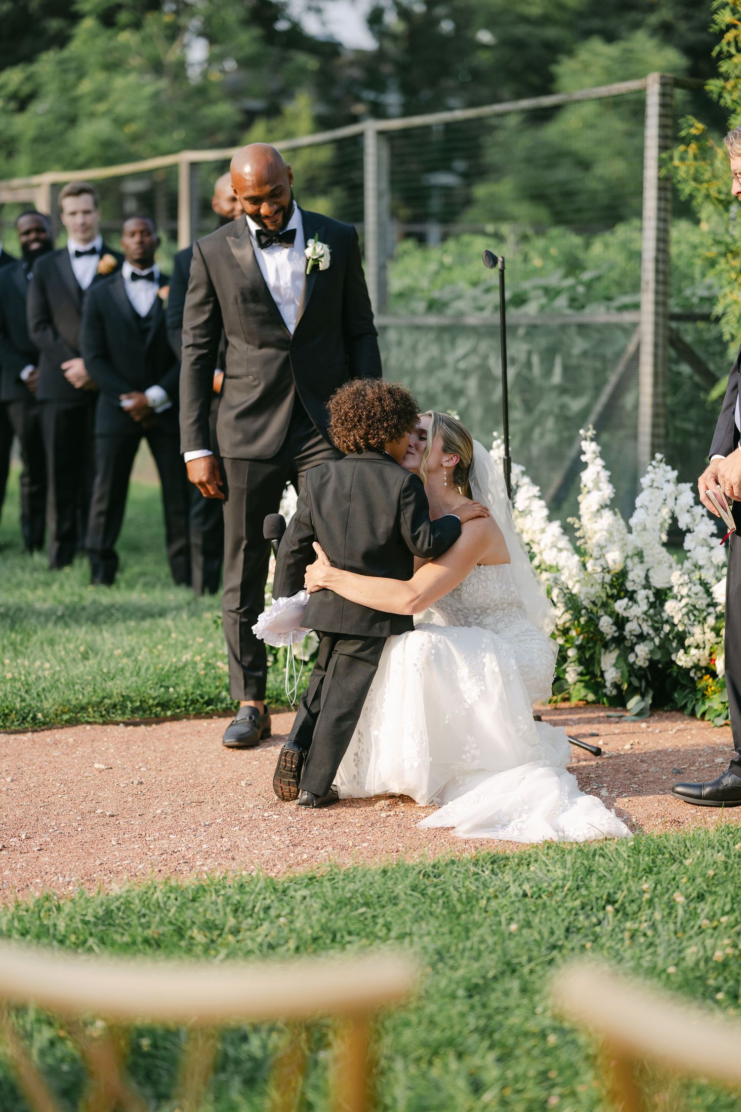 A tender moment during an outdoor wedding ceremony with attendants standing in the background on a grassy lawn. - Elawa Farm