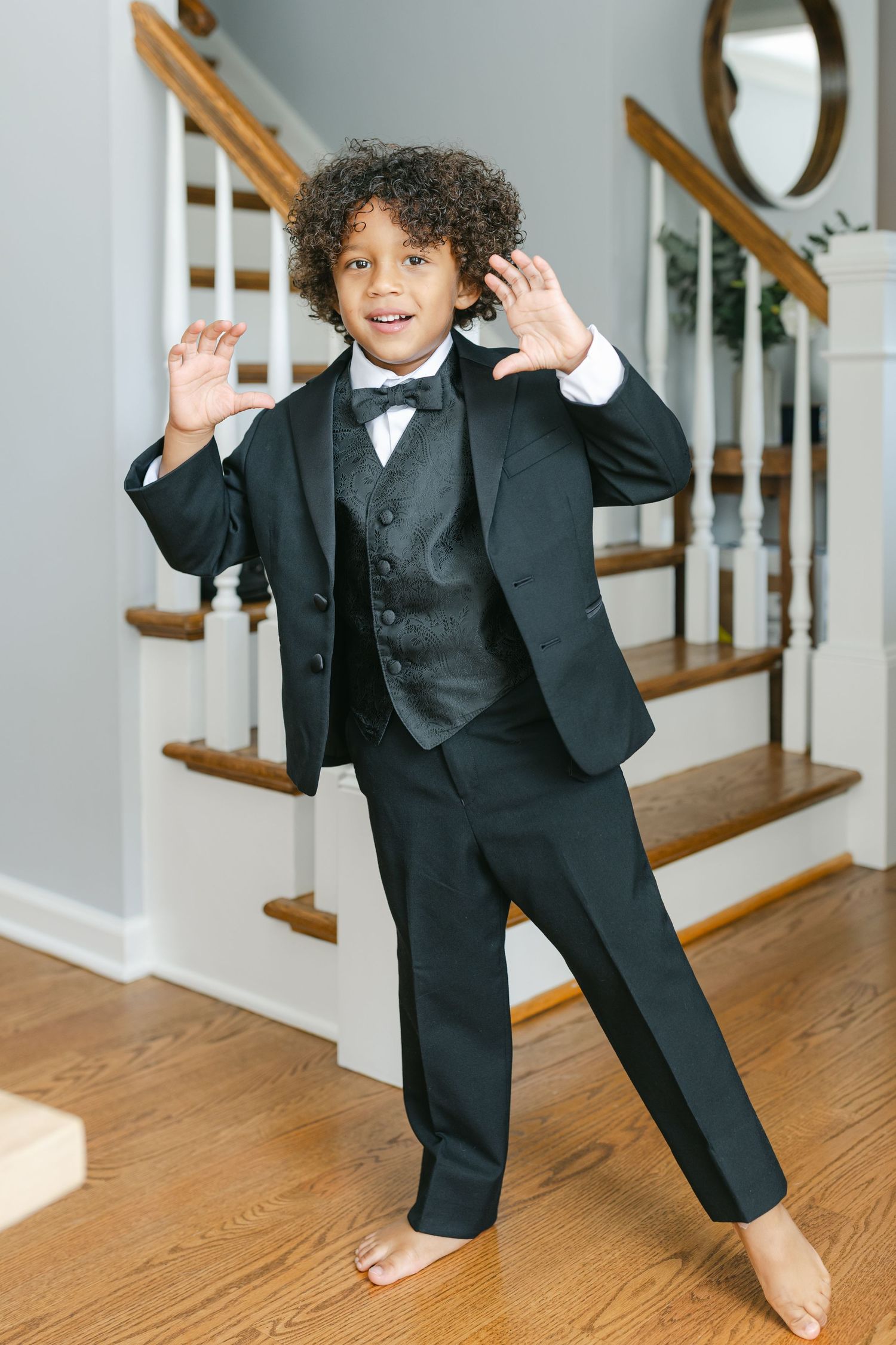 Two people in formal attire stand in a hallway near wooden stairs as one helps adjust the other's outfit.