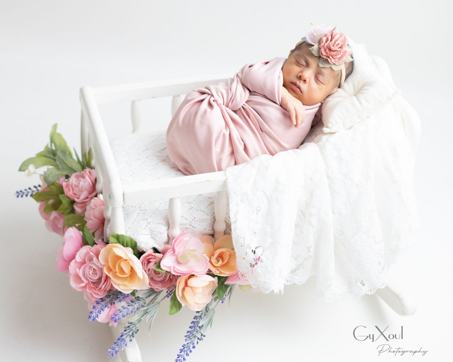 A newborn baby sleeps peacefully in a white basket decorated with pink and peach roses and lavender flowers.