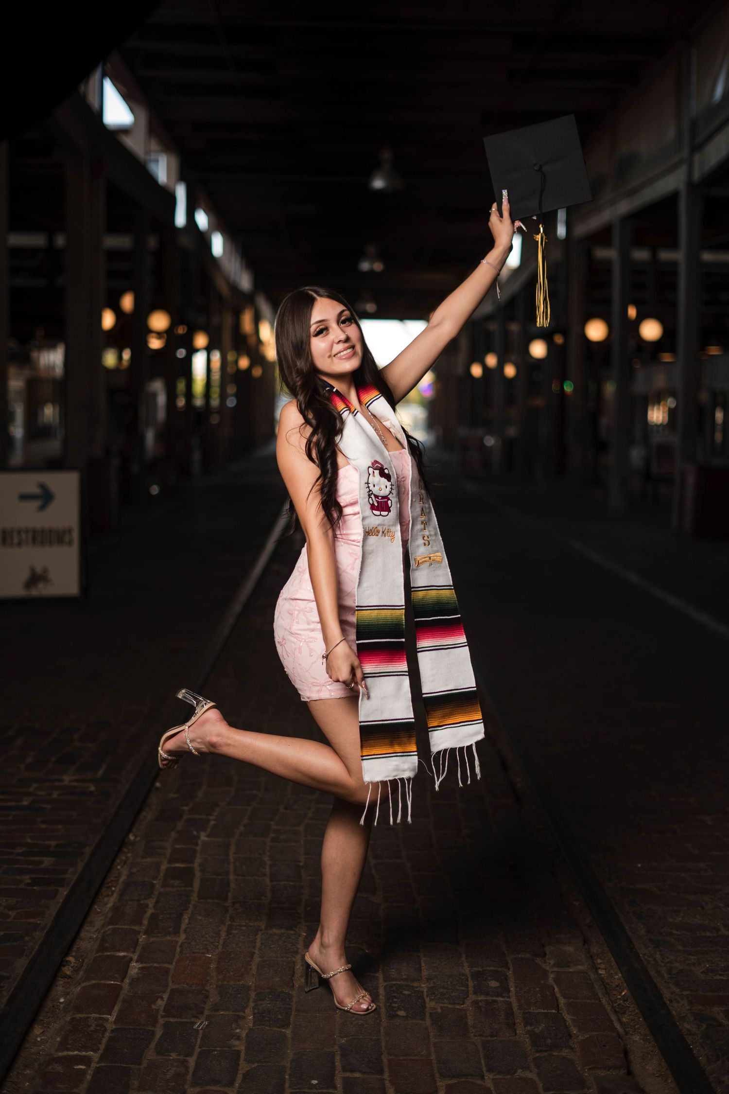 Female graduate celebrates in pink dress holding cap aloft wearing colorful serape stole at night.