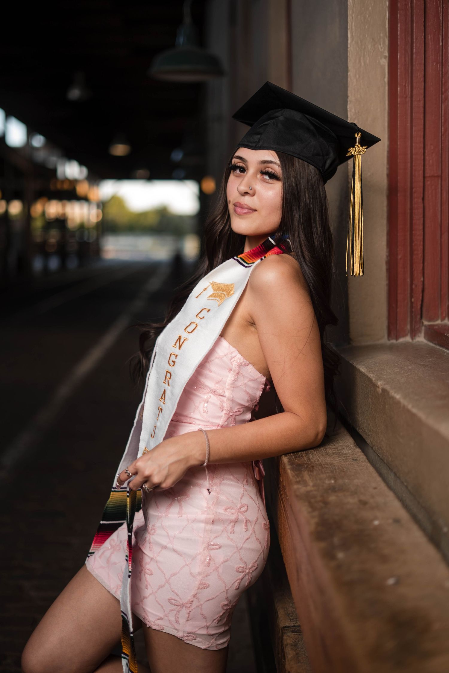 Smiling female graduate in pink dress and black cap leans against wall wearing congratulations sash.