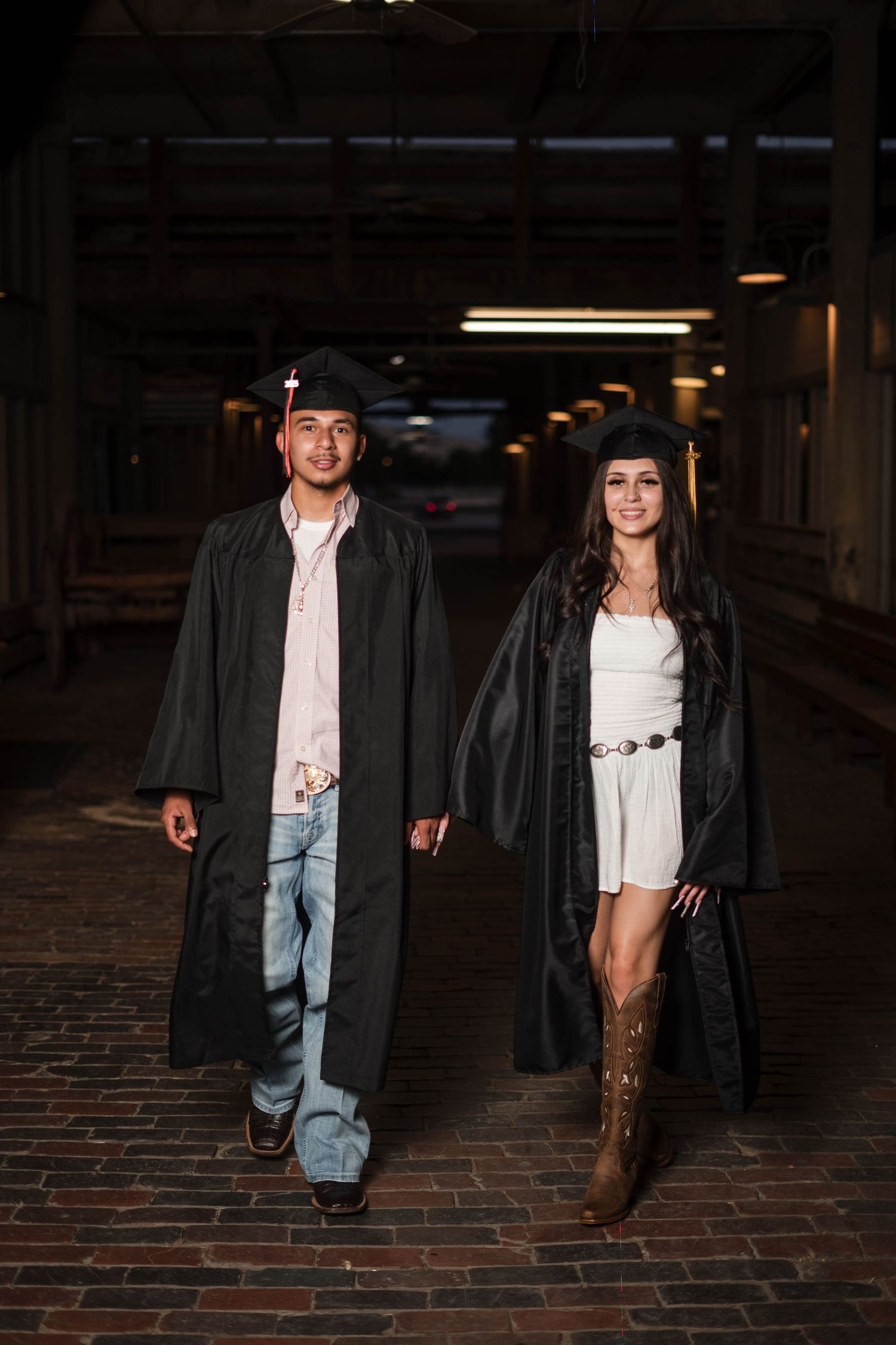 Two graduates in black caps and gowns walk together through a brick corridor at night.
