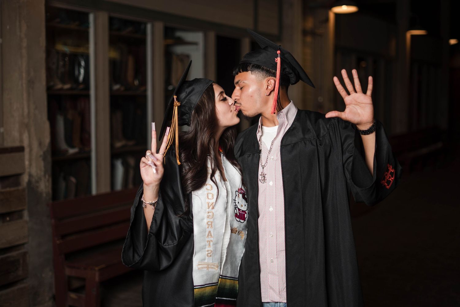 Two graduates in black gowns share a kiss while holding diplomas outside at night.