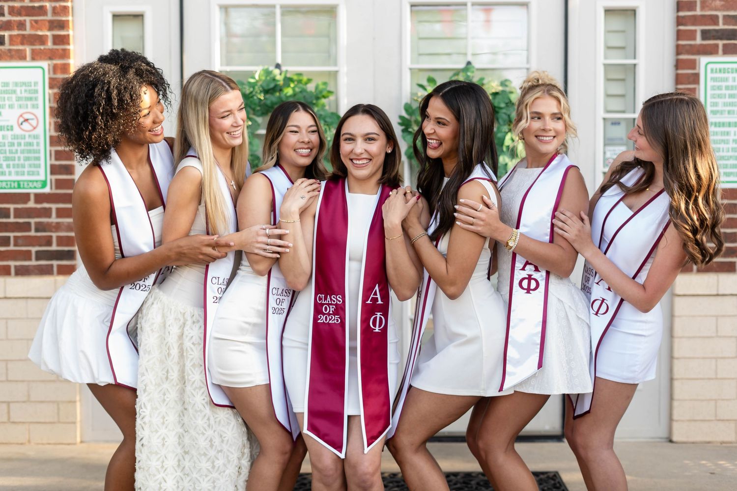 Group of seven smiling Alpha Phi sorority women celebrate graduation in white dresses outdoors.