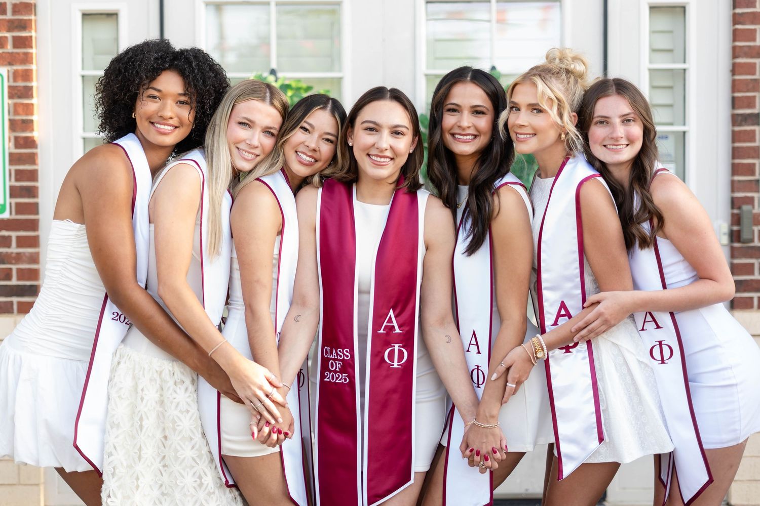 Seven college women in white dresses pose together wearing Alpha Phi graduation stoles.
