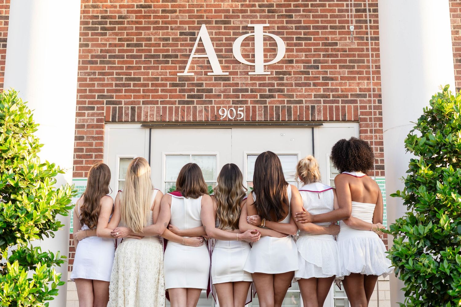 Seven women in white dresses pose with arms around each other in front of Alpha Phi sorority house number 905.