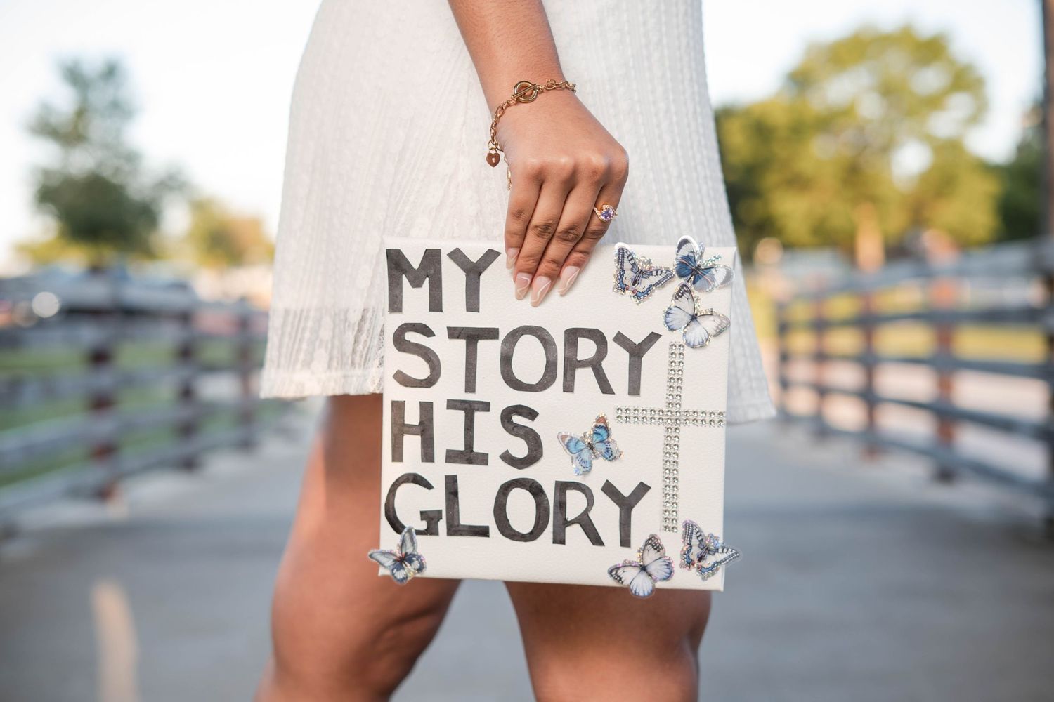 Woman holding a decorative sign reading My Story His Glory adorned with butterfly designs outdoors.