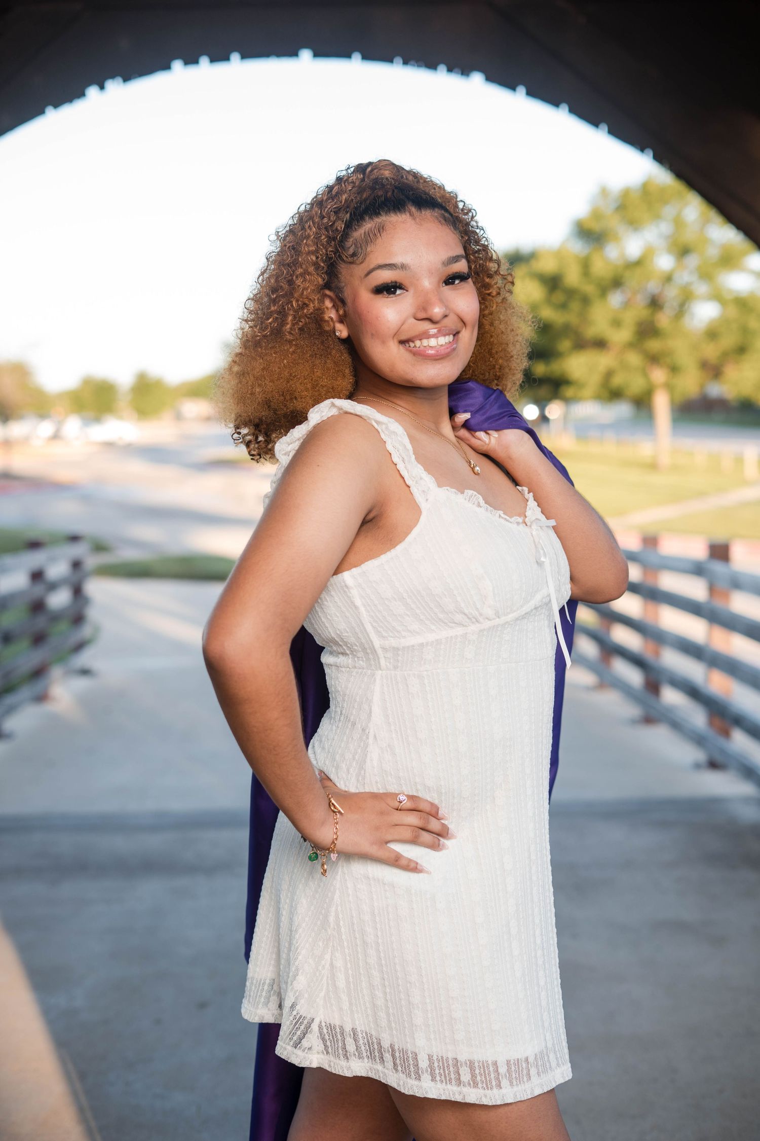Smiling young woman in white dress holding purple graduation gown under an arch.