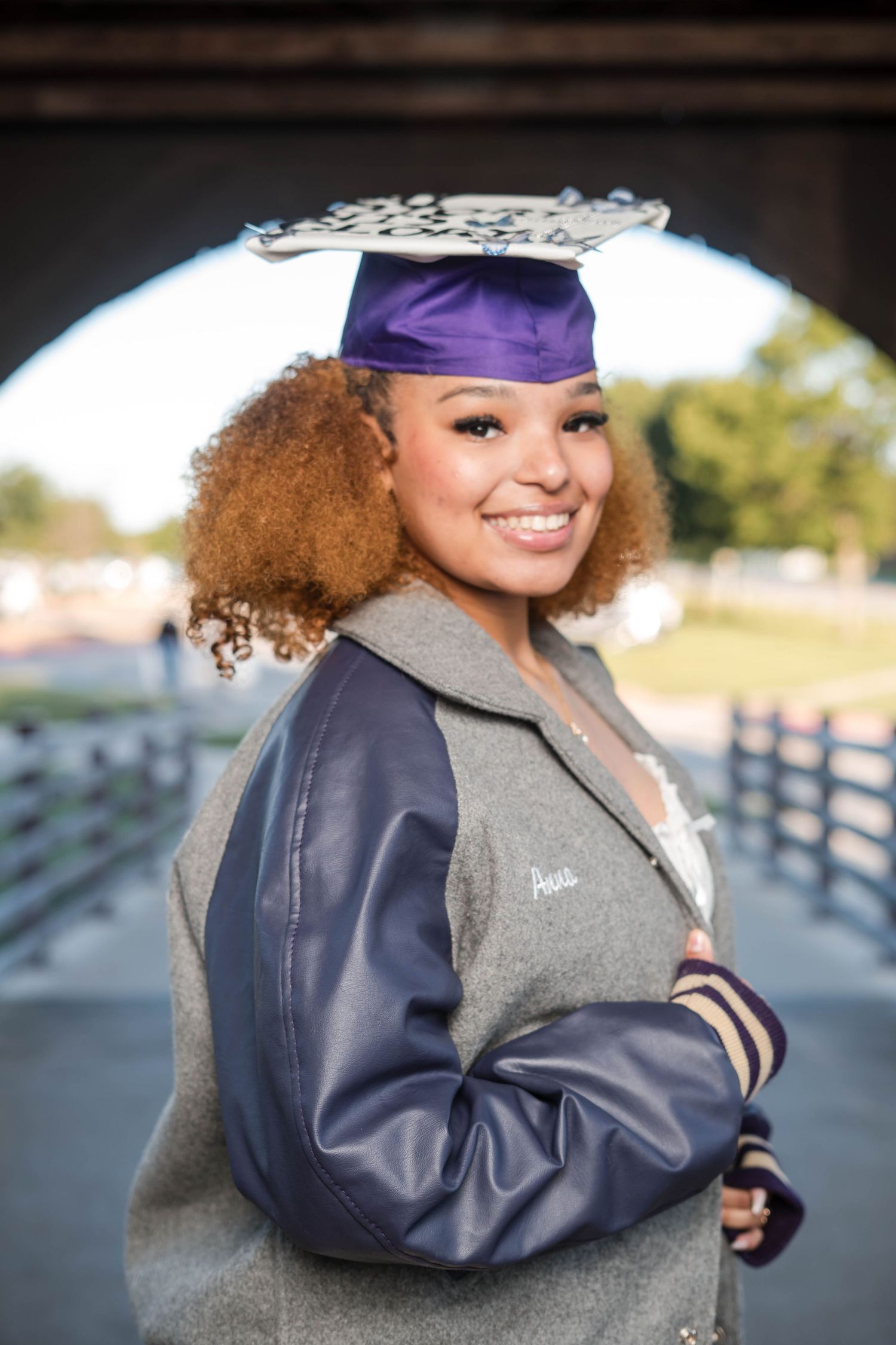 Smiling graduate wearing purple cap and varsity jacket posing under a bridge arch outdoors.