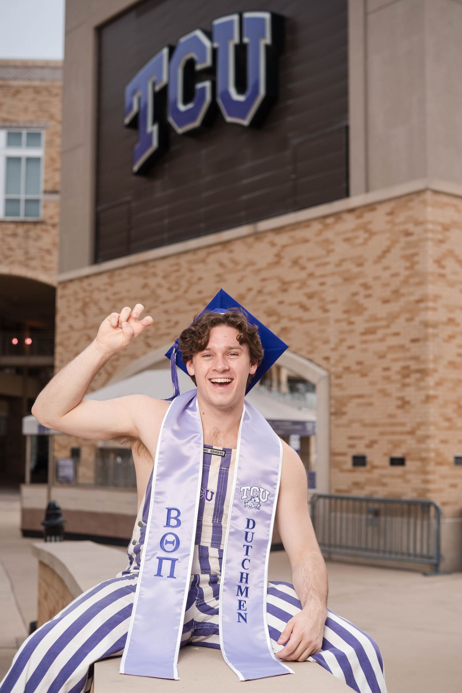 Happy male TCU graduate in blue cap sits outdoors wearing Beta Theta Pi and Dutchmen stoles.