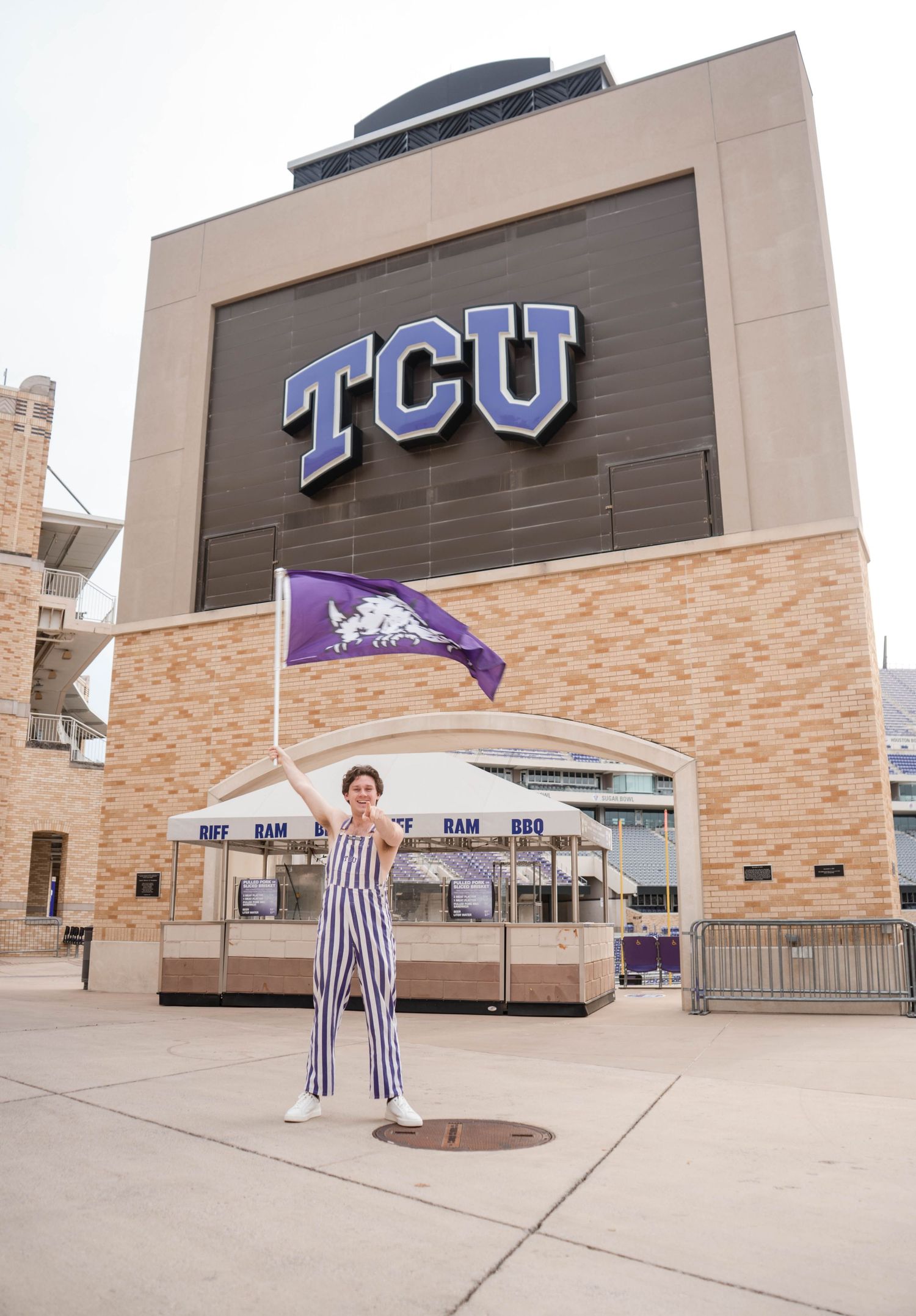 Excited graduate waves purple TCU Horned Frogs flag in striped jumpsuit outside stadium.