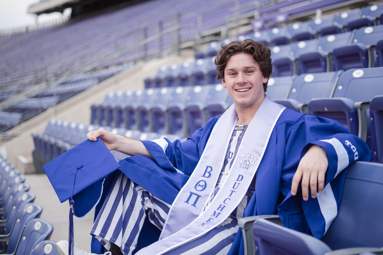 Male TCU graduate in blue cap and gown relaxes in stadium seats wearing fraternity stole.