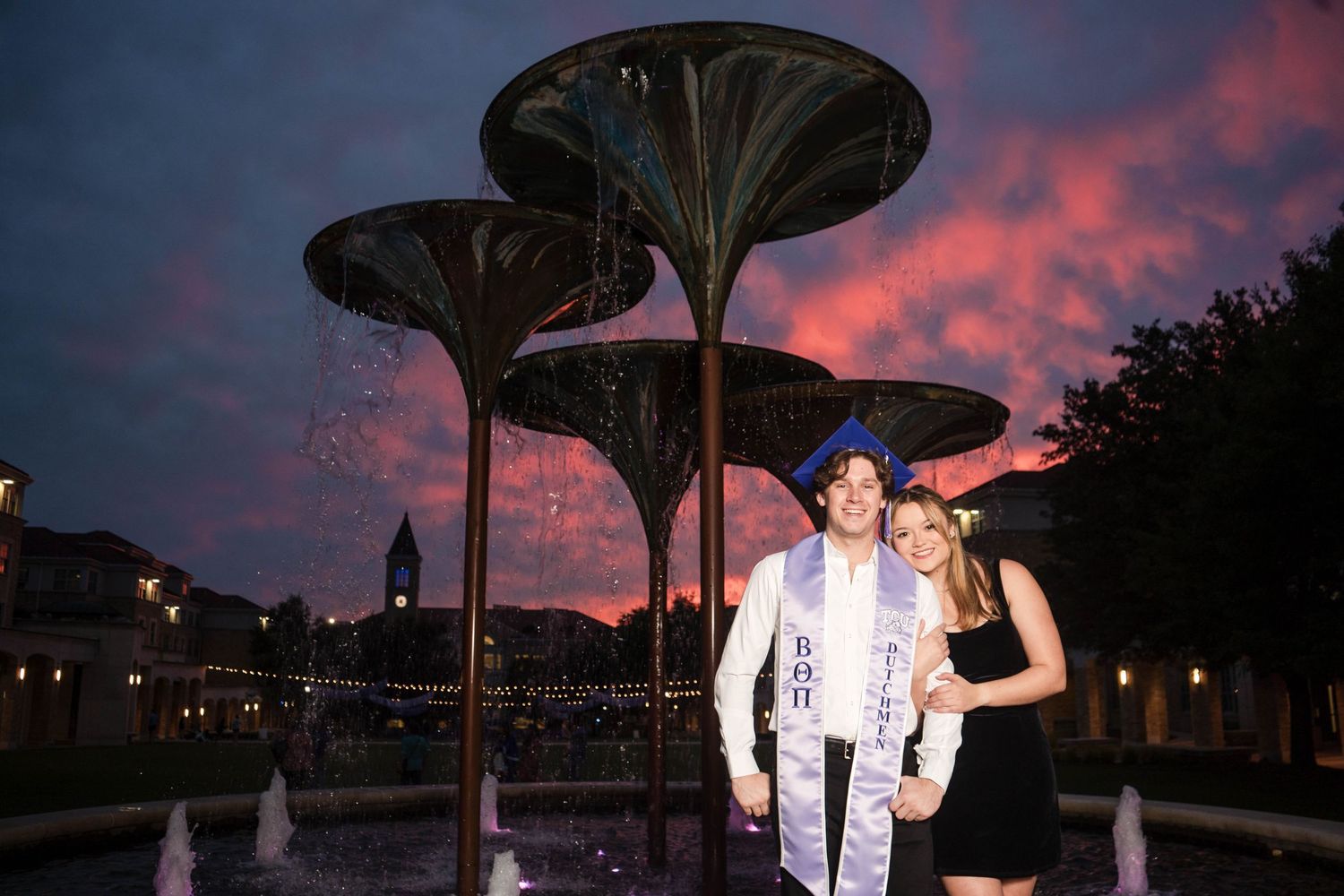 Couple poses together at sunset near decorative fountain with dramatic pink and purple sky.