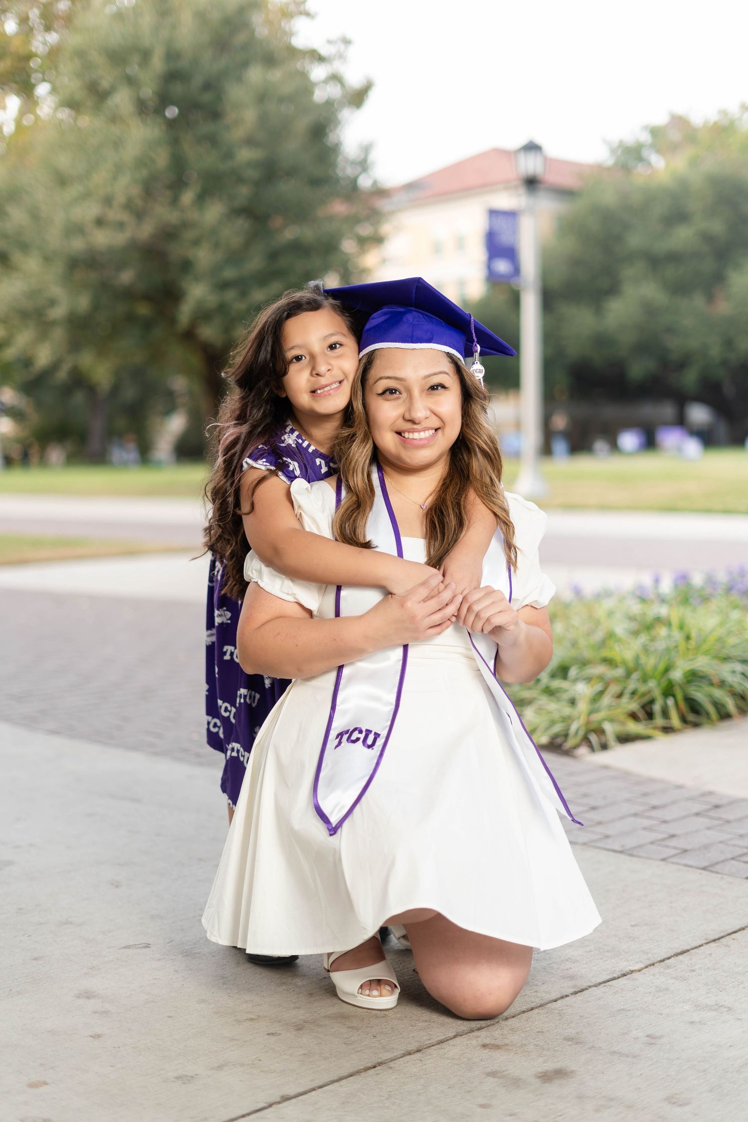 TCU graduate in blue cap and white dress posing with young girl on campus.