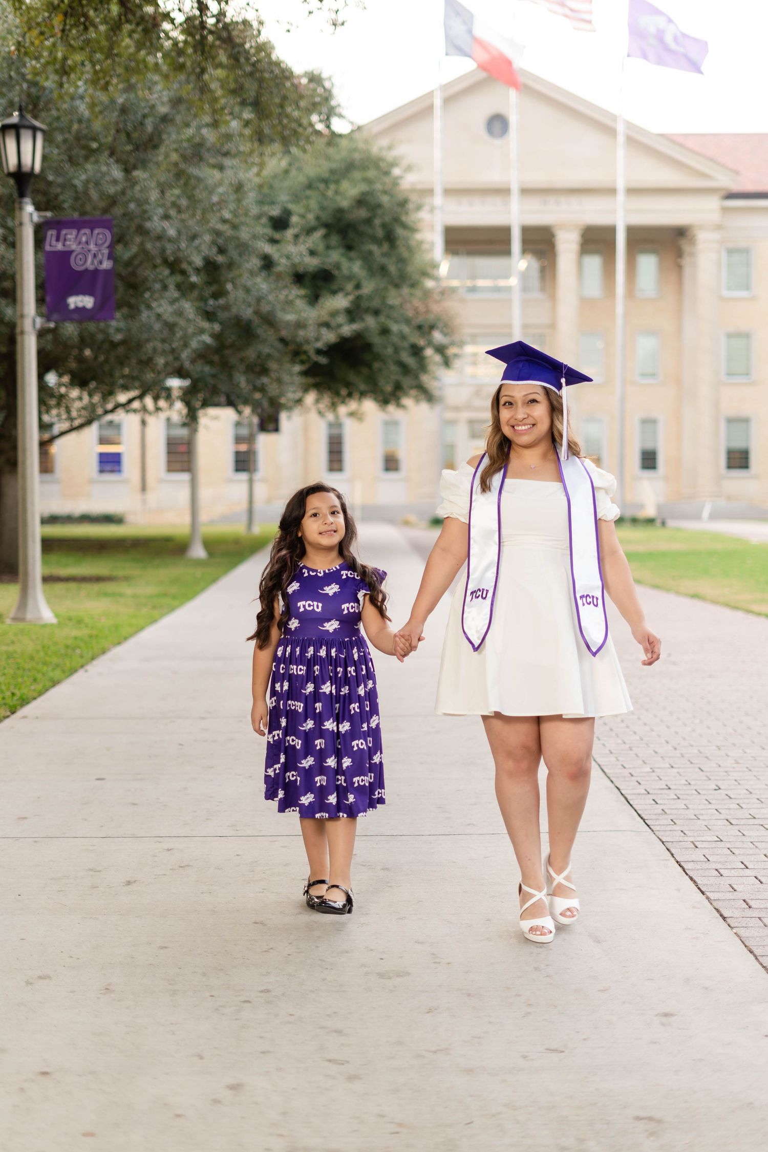 TCU graduate in blue cap and white dress walking hand-in-hand with young girl on campus.