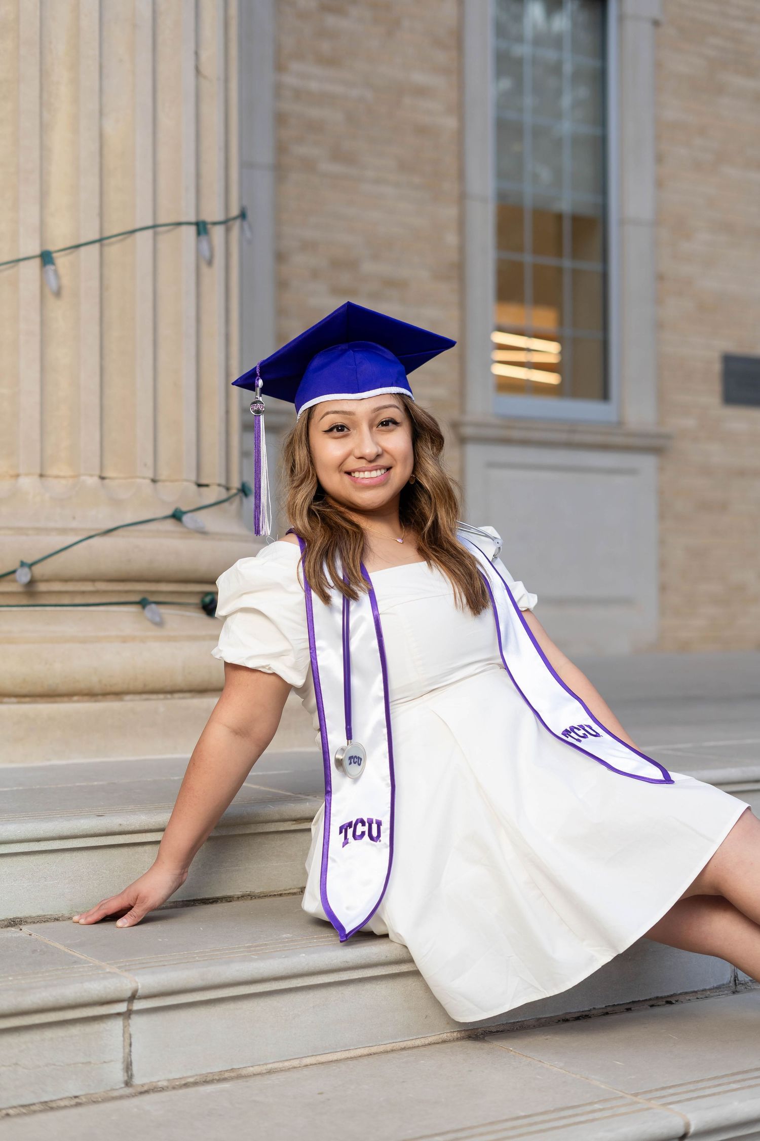 Smiling TCU graduate in blue cap and white dress sitting on university building steps.