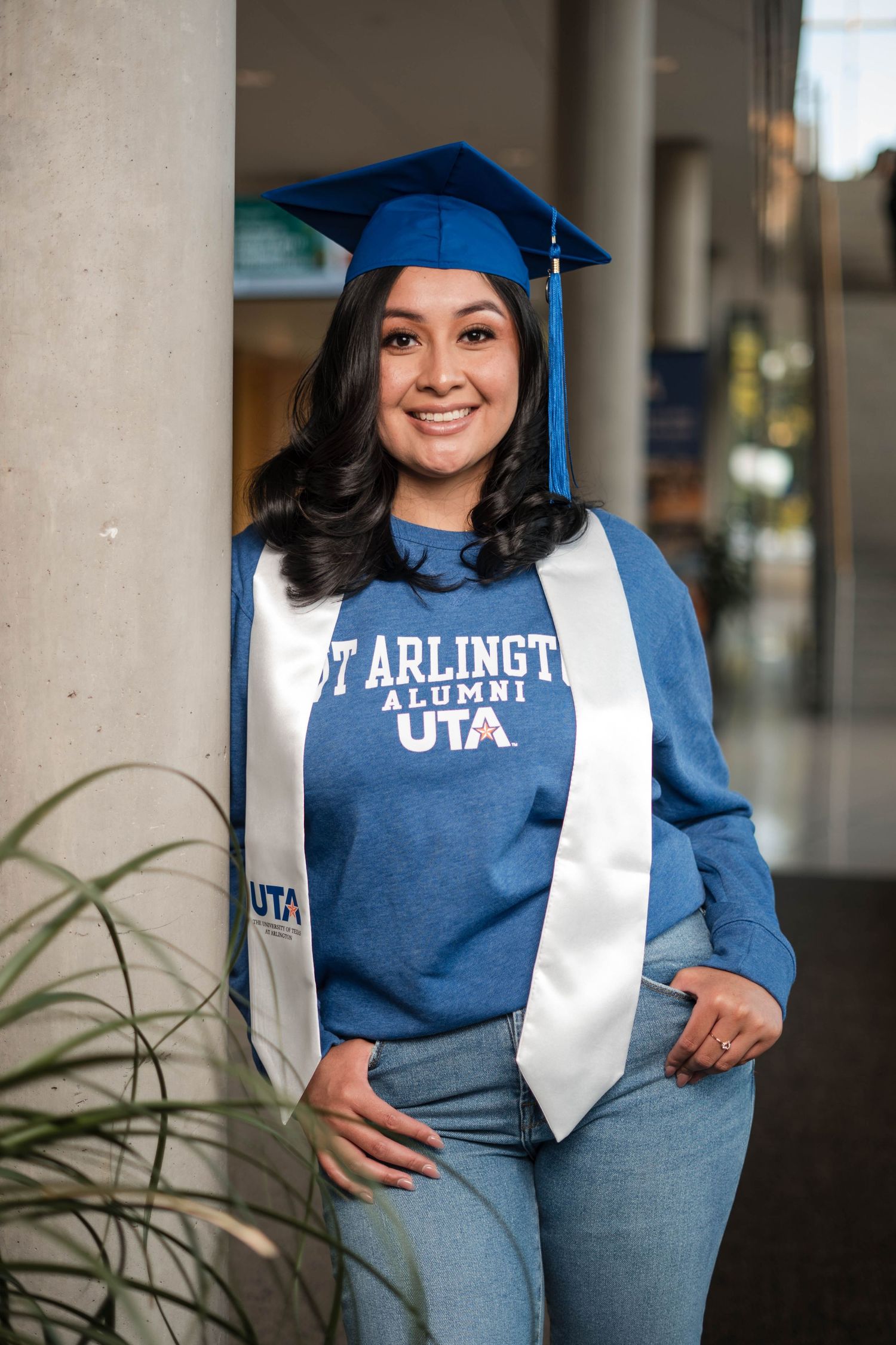 Female UTA graduate smiling in blue cap and Arlington Alumni sweatshirt with white stole.