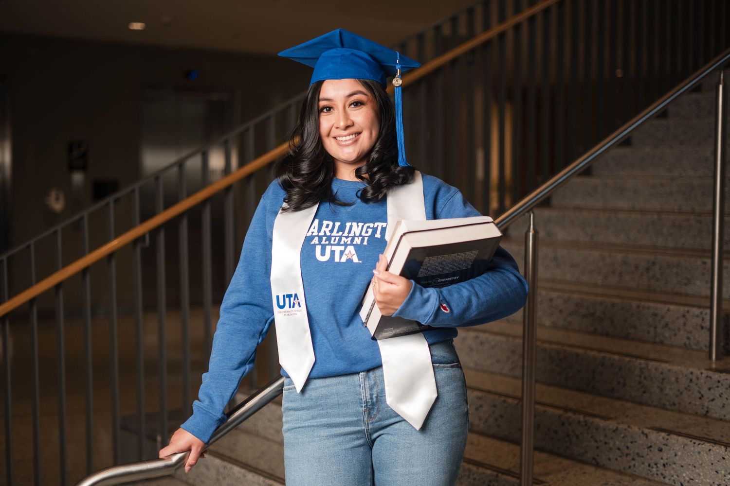 Female UTA graduate in blue cap holding books on university staircase.