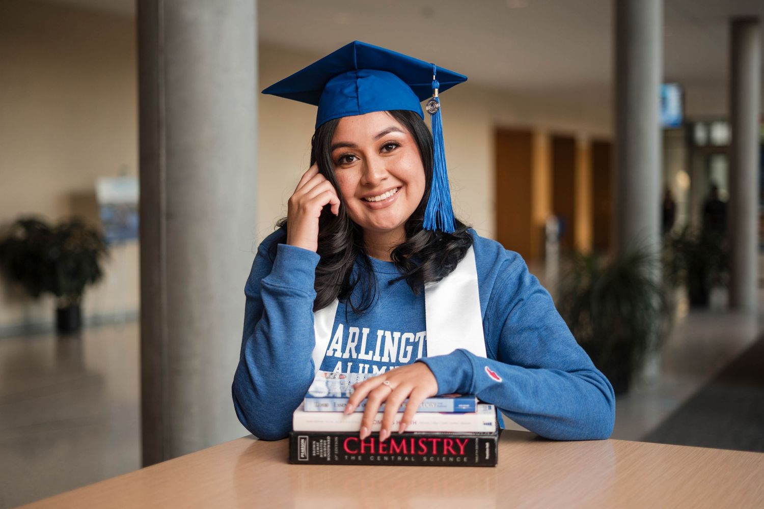 Smiling female graduate in blue cap and UTA sweatshirt with chemistry textbooks.