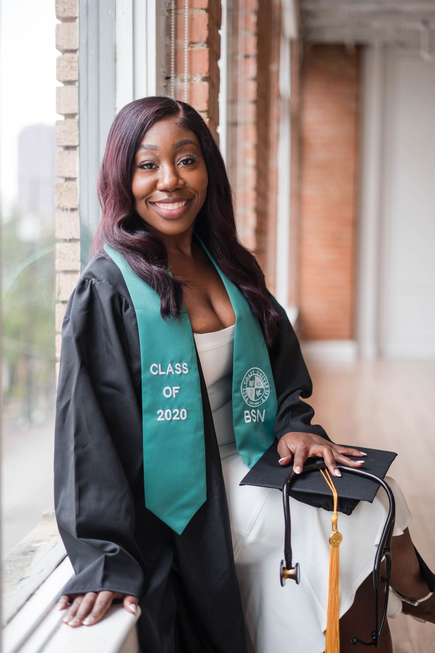 Female nursing graduate in black gown and green BSN stole poses by window with cap.