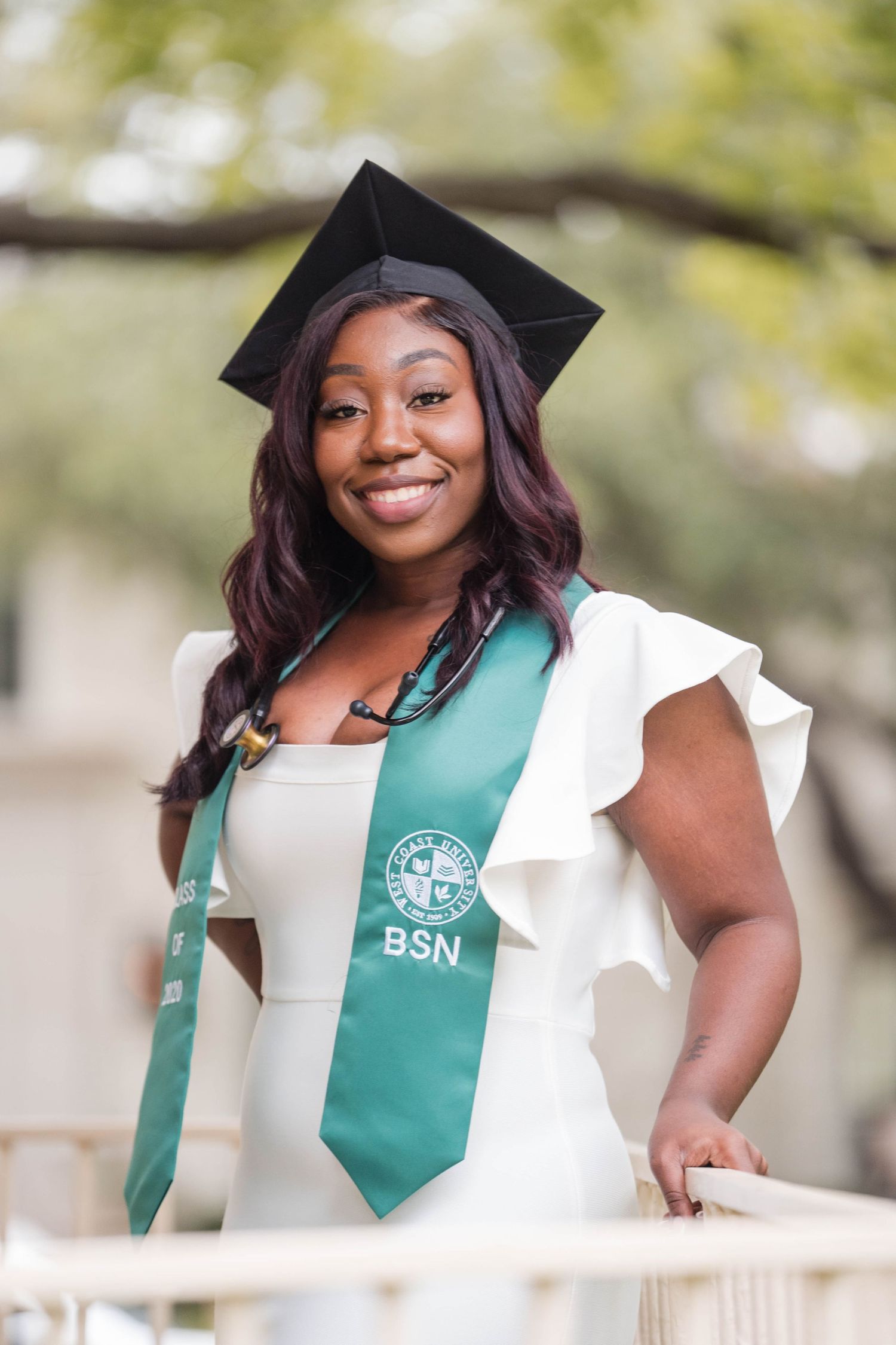 Smiling woman in white dress wears black cap and green BSN graduation stole outdoors.