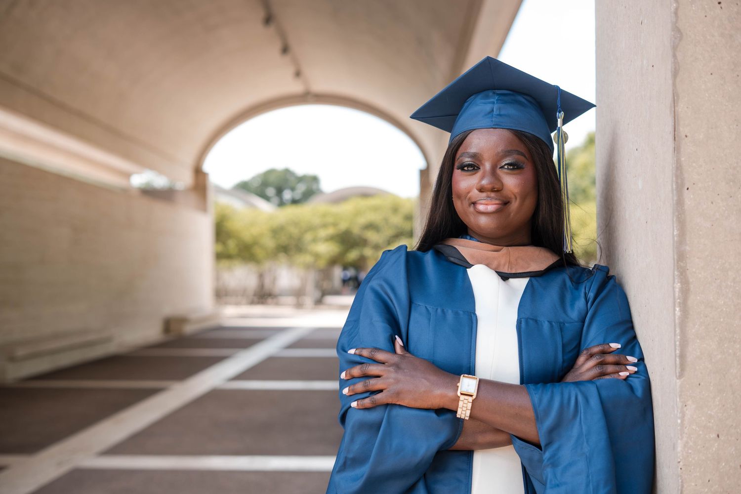 Smiling Black woman in blue graduation cap and gown poses with arms crossed under an archway outdoors.