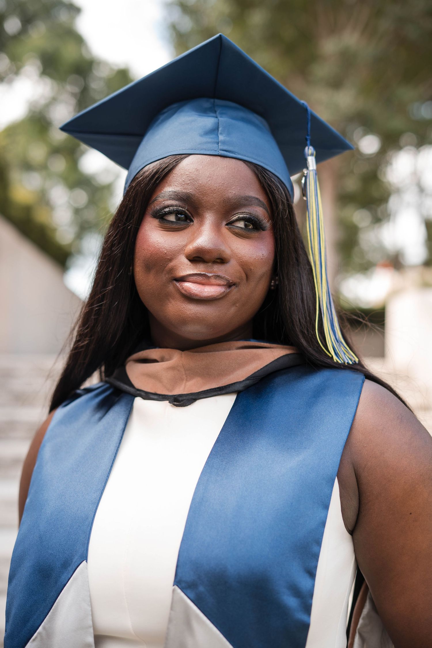 Smiling young woman in blue graduation cap and gown posing outdoors for her graduation photo.