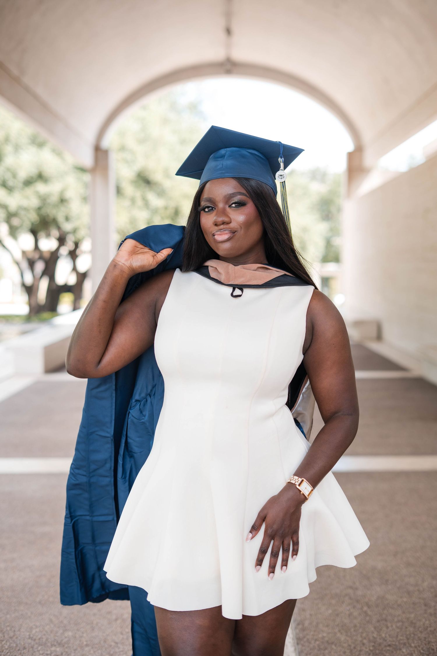 Young Black woman in white dress and navy graduation cap and gown smiles proudly at her college graduation.