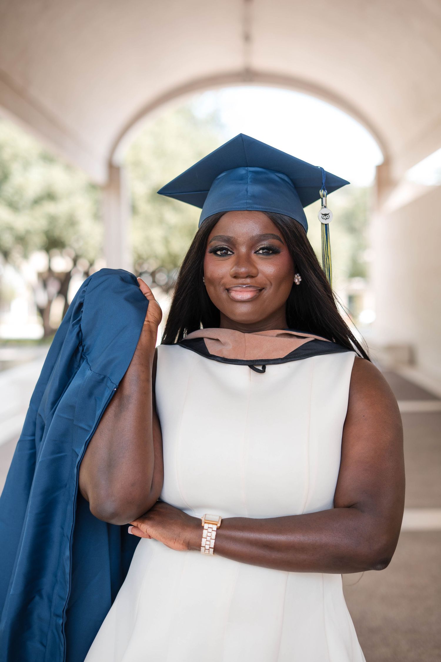 Smiling graduate in navy cap and white dress holds gown over shoulder under arched walkway.
