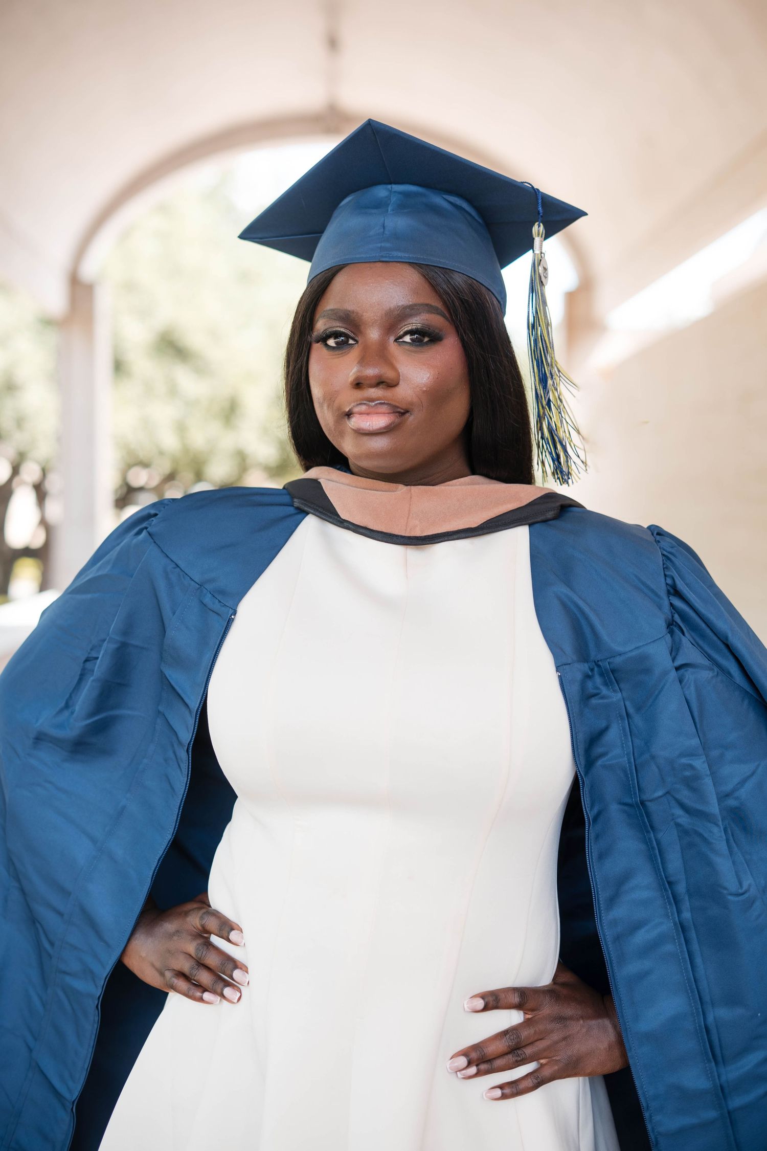Young woman in blue graduation cap and gown over white dress posing outdoors at university campus.