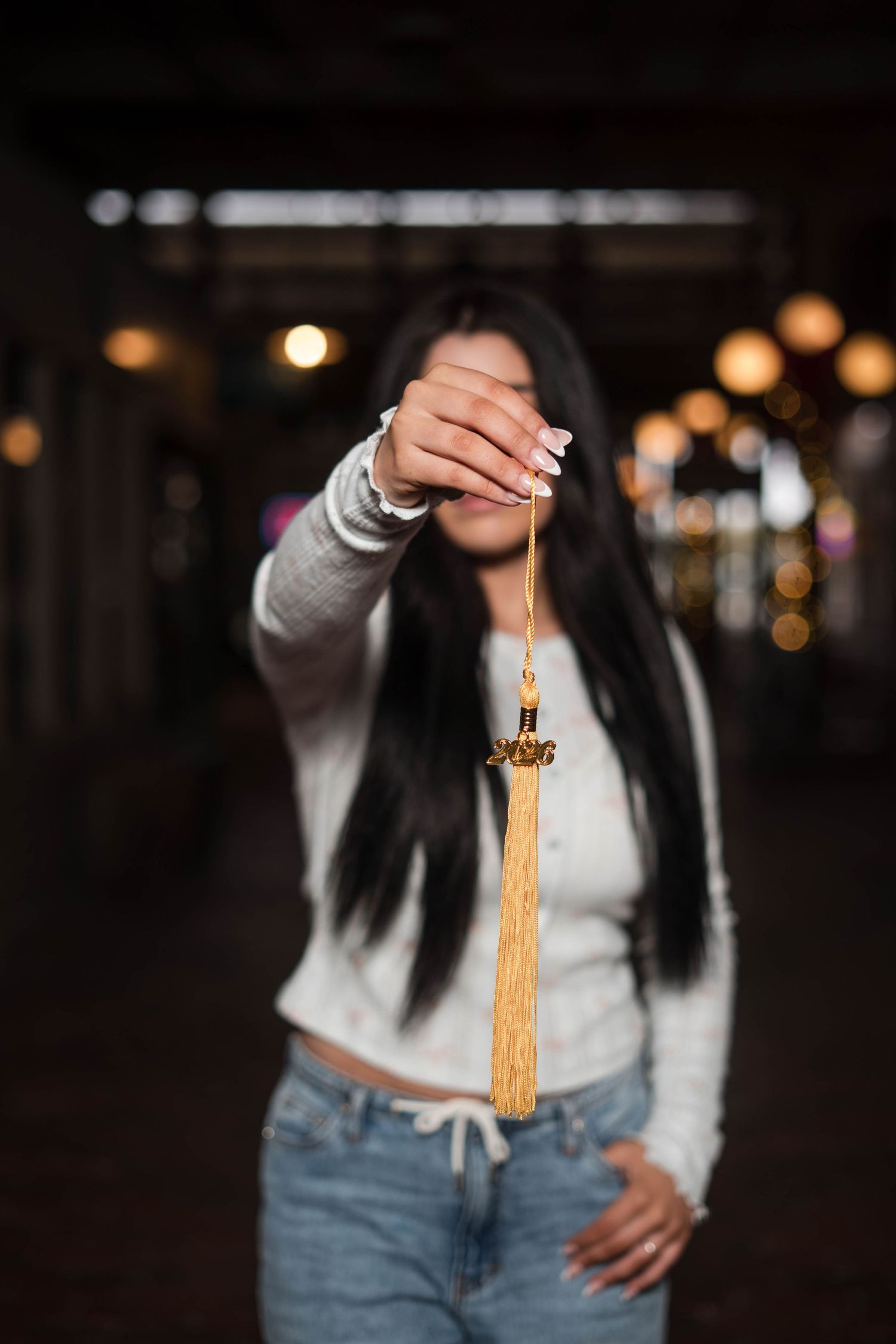 Young woman holding a gold tassel toward the camera in a dimly lit venue.