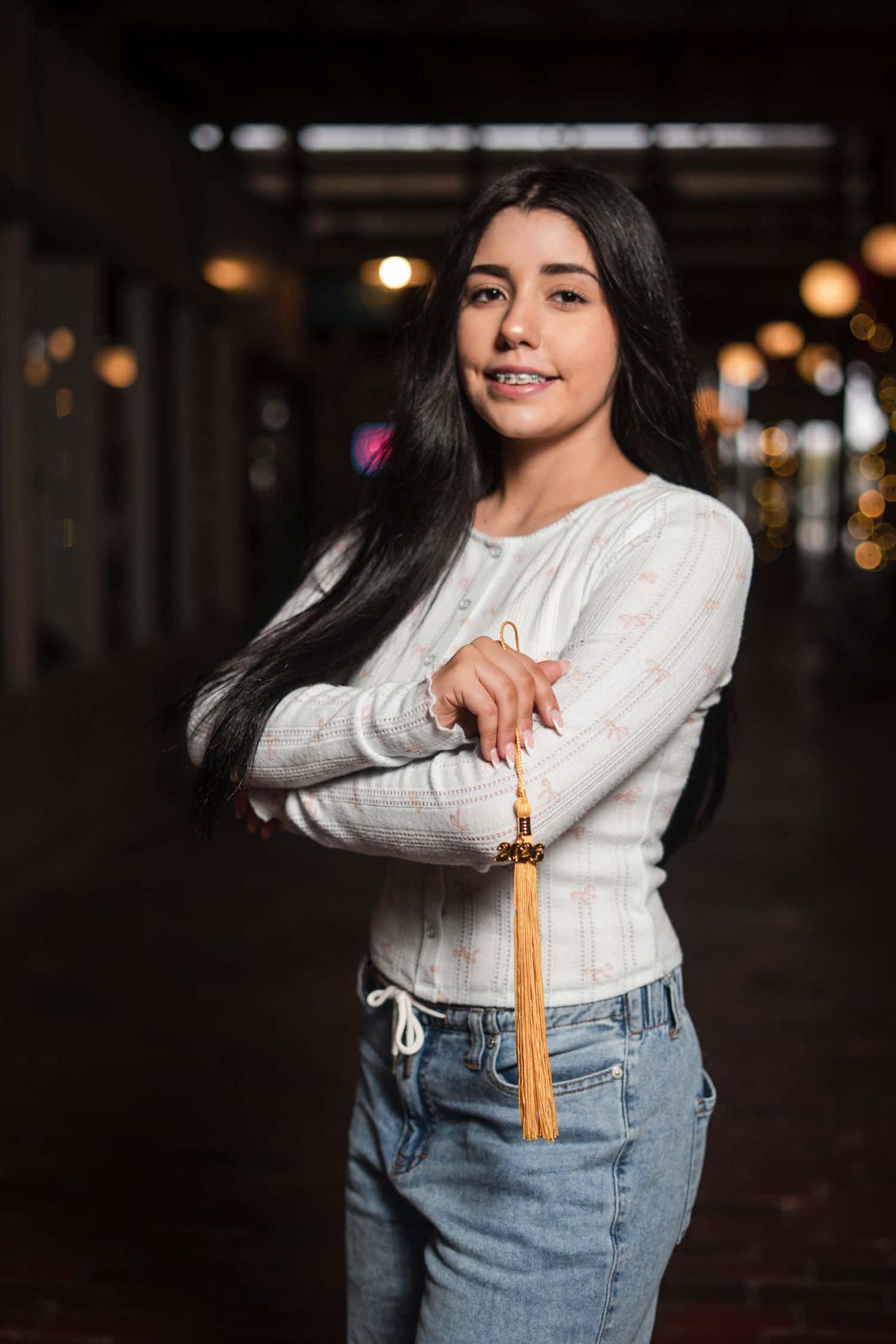 Smiling young woman with long dark hair holding a gold tassel in a dark hallway.