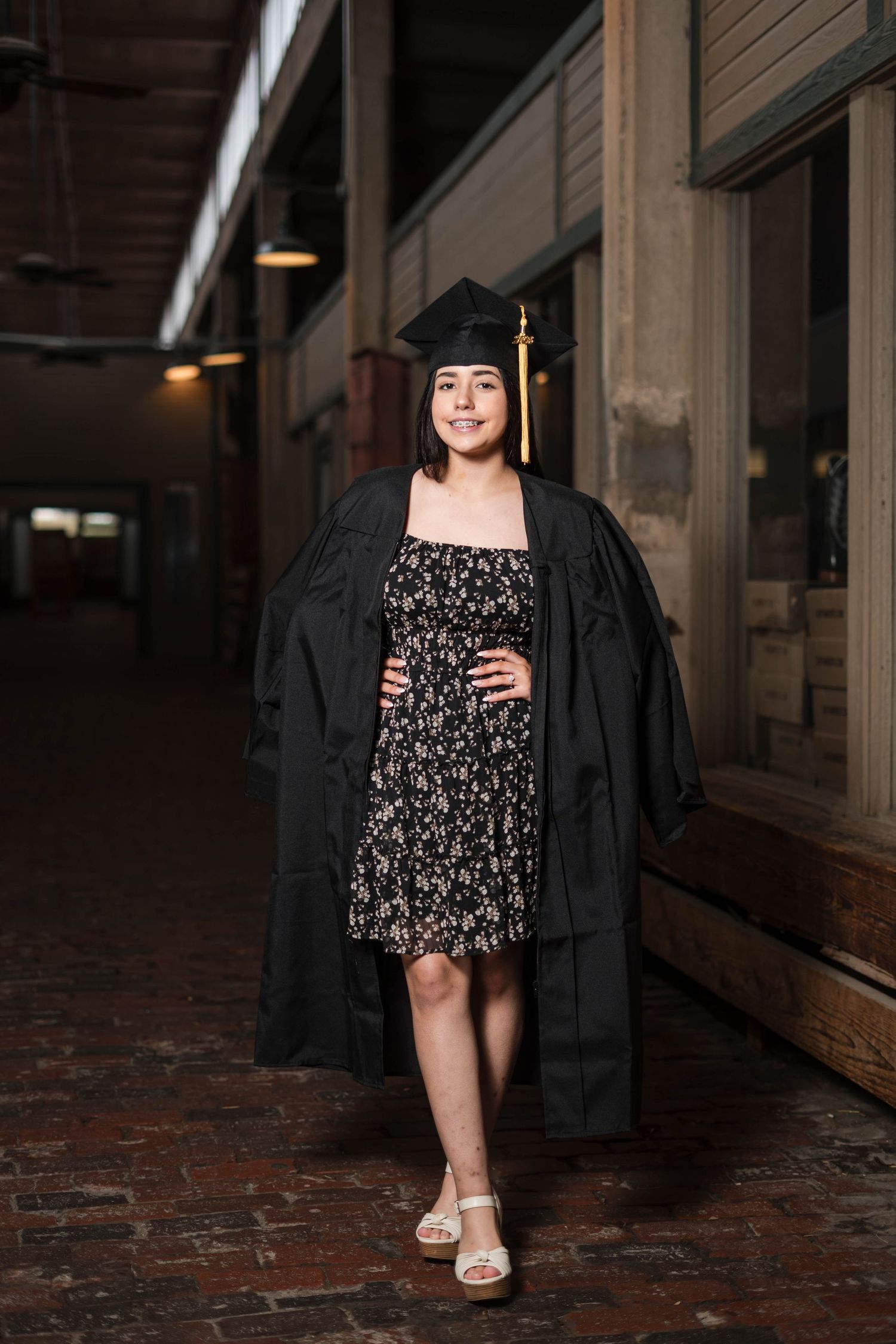 Smiling graduate in cap and gown over floral dress walking in an indoor corridor.