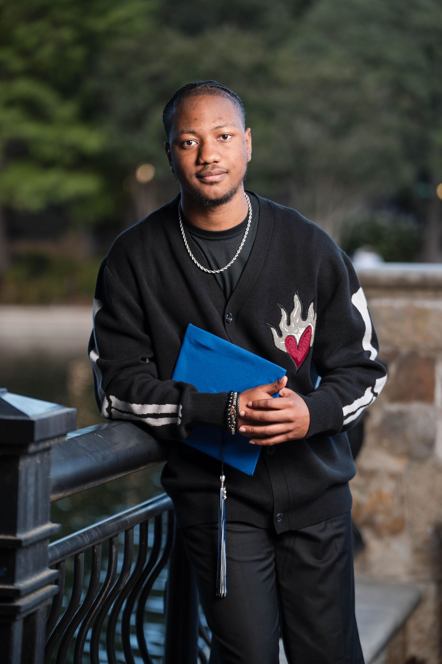Young man in black graphic jacket holds blue folder while leaning on iron railing outdoors.