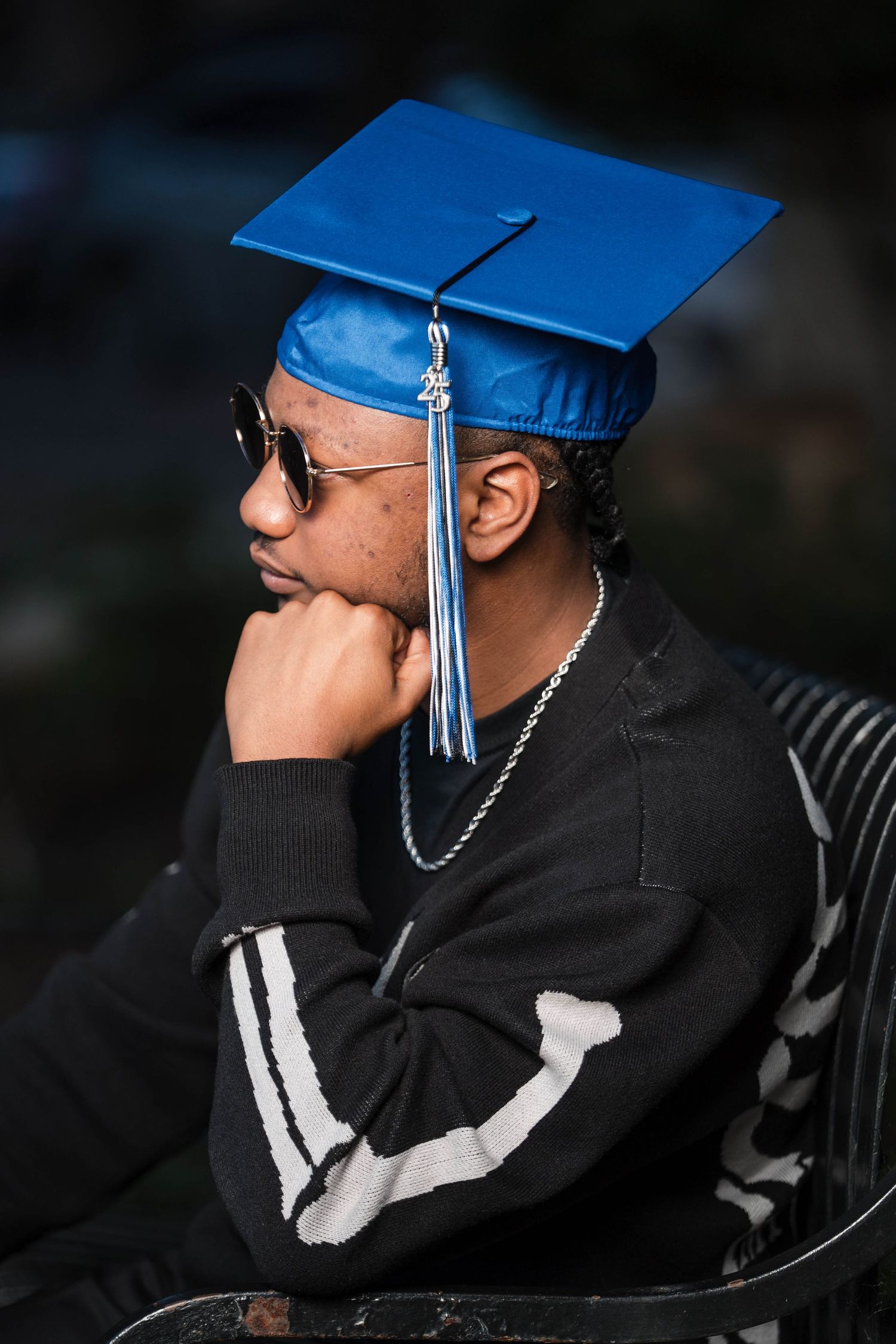 Young male graduate wearing blue cap and sunglasses sits thoughtfully on bench outdoors.